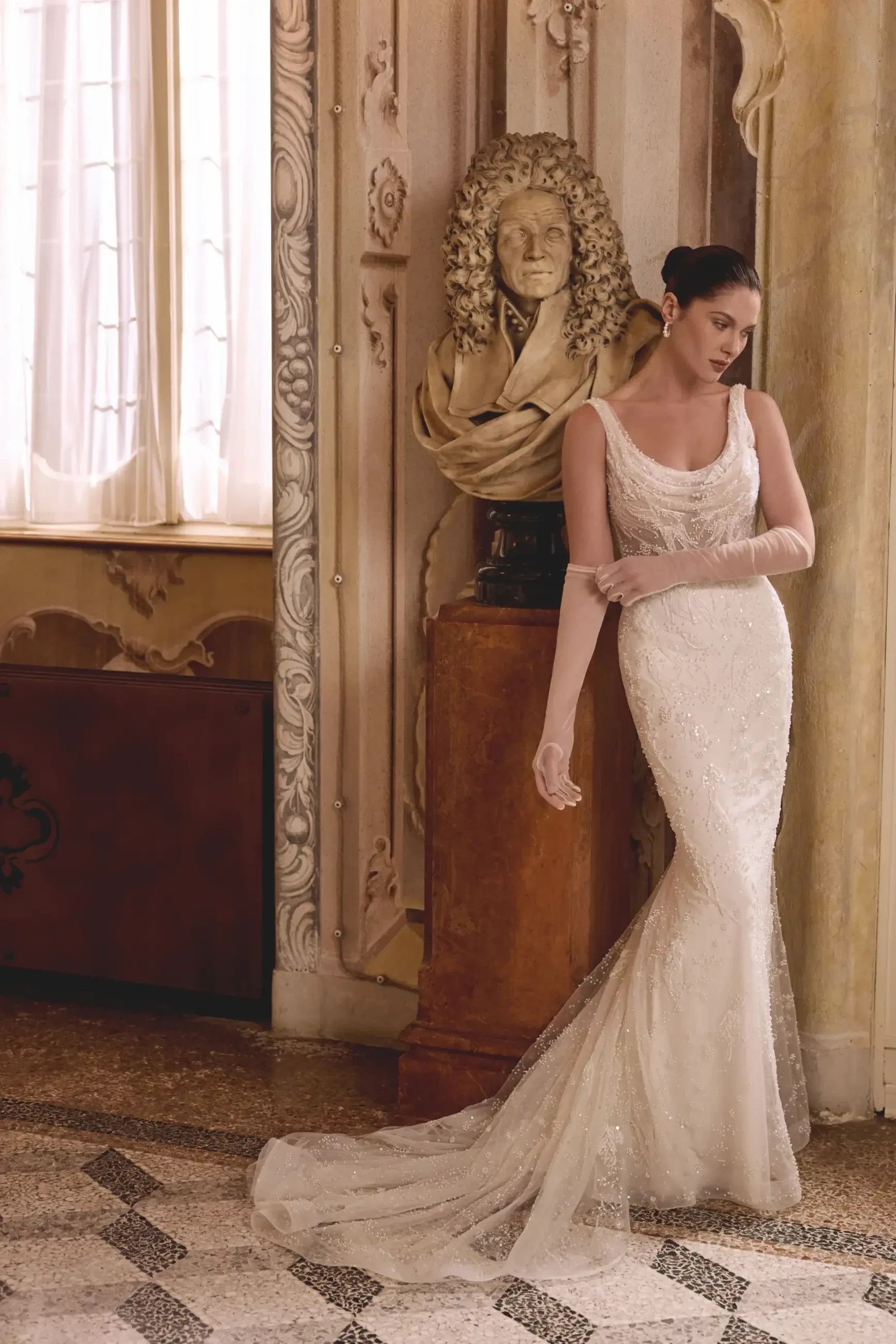 A woman in a glamorous wedding dress stands beside a sculpture in an elegant interior space. The dress features intricate detailing, and she poses gracefully with one arm resting on the sculpture's base.