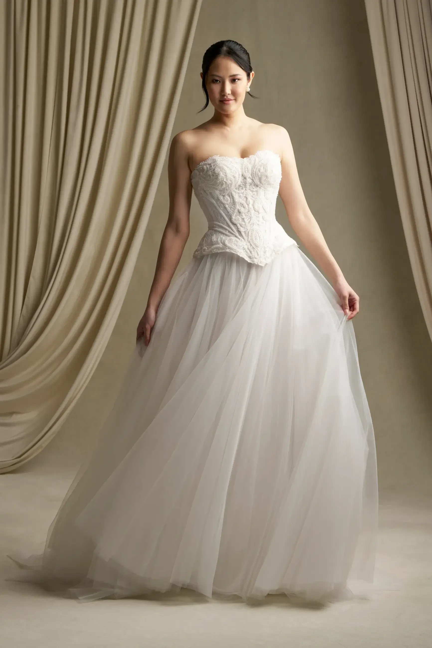 A woman wearing a strapless, white wedding gown standing in front of a draped backdrop.