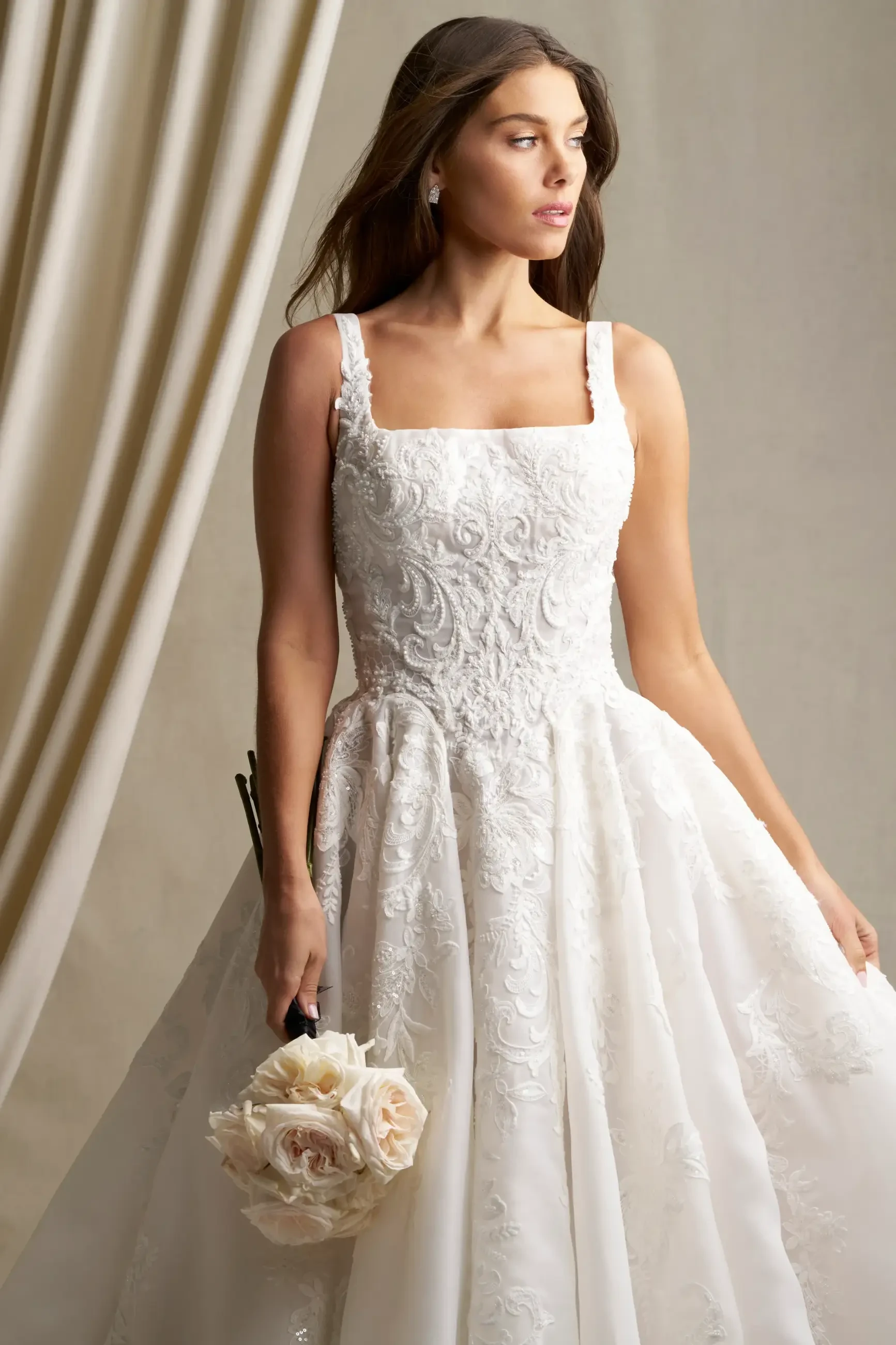 A woman in a white wedding dress with a floral bouquet, standing gracefully against a soft, neutral background.