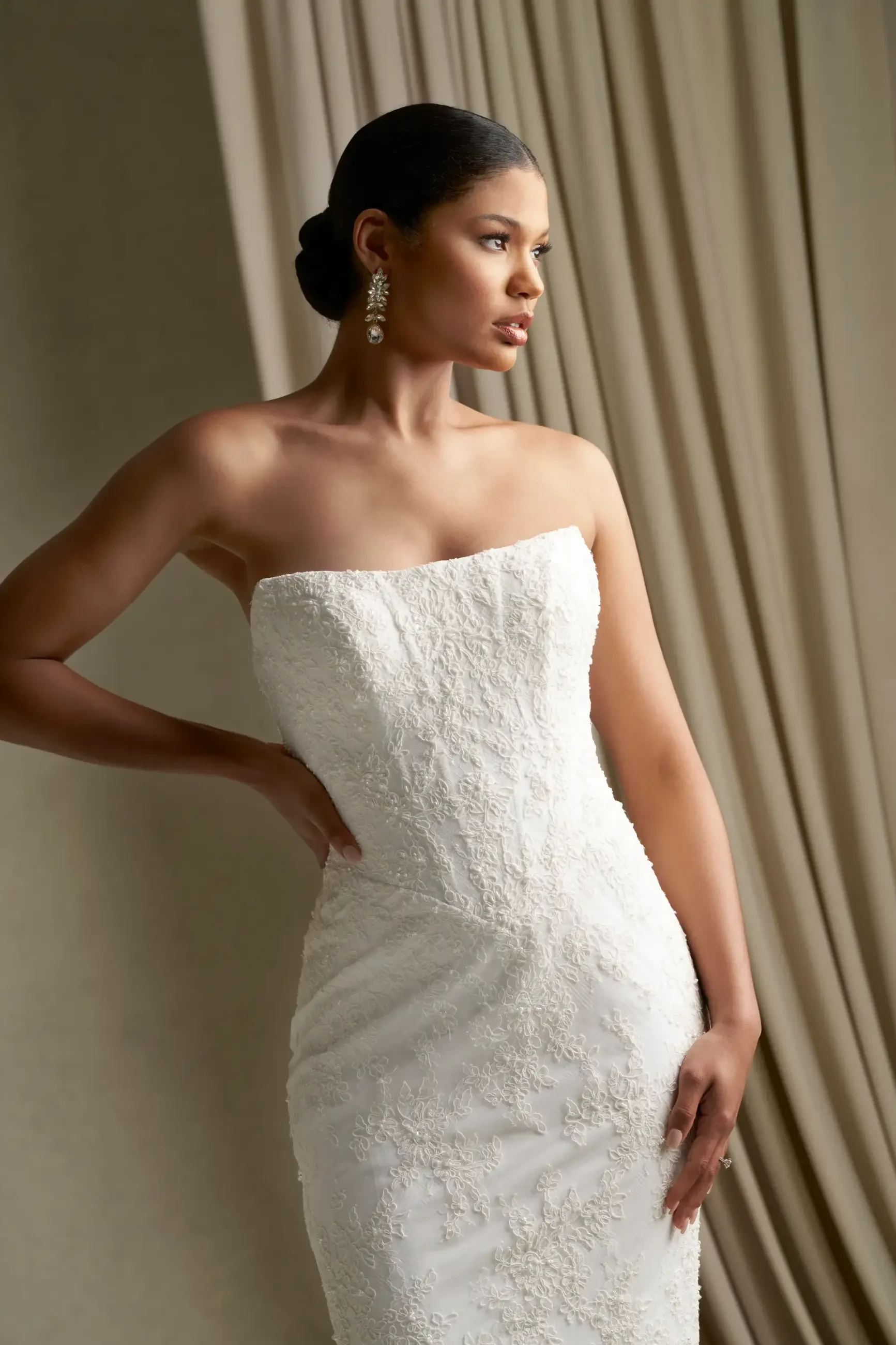A model wearing a strapless white wedding dress with intricate embroidery, standing elegantly with one hand on her hip and the other resting by her side.